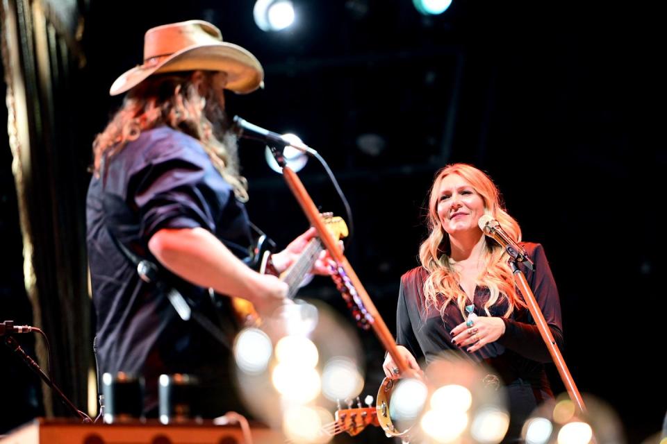 chris stapleton plays guitar and faces away from the camera, morgane stapleton faces him, they stand on a stage each in front of microphone stands