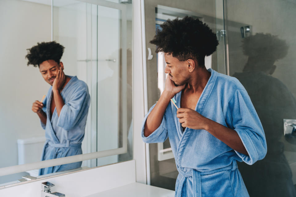 A person in a blue robe brushes their teeth in front of a bathroom mirror while touching their cheek