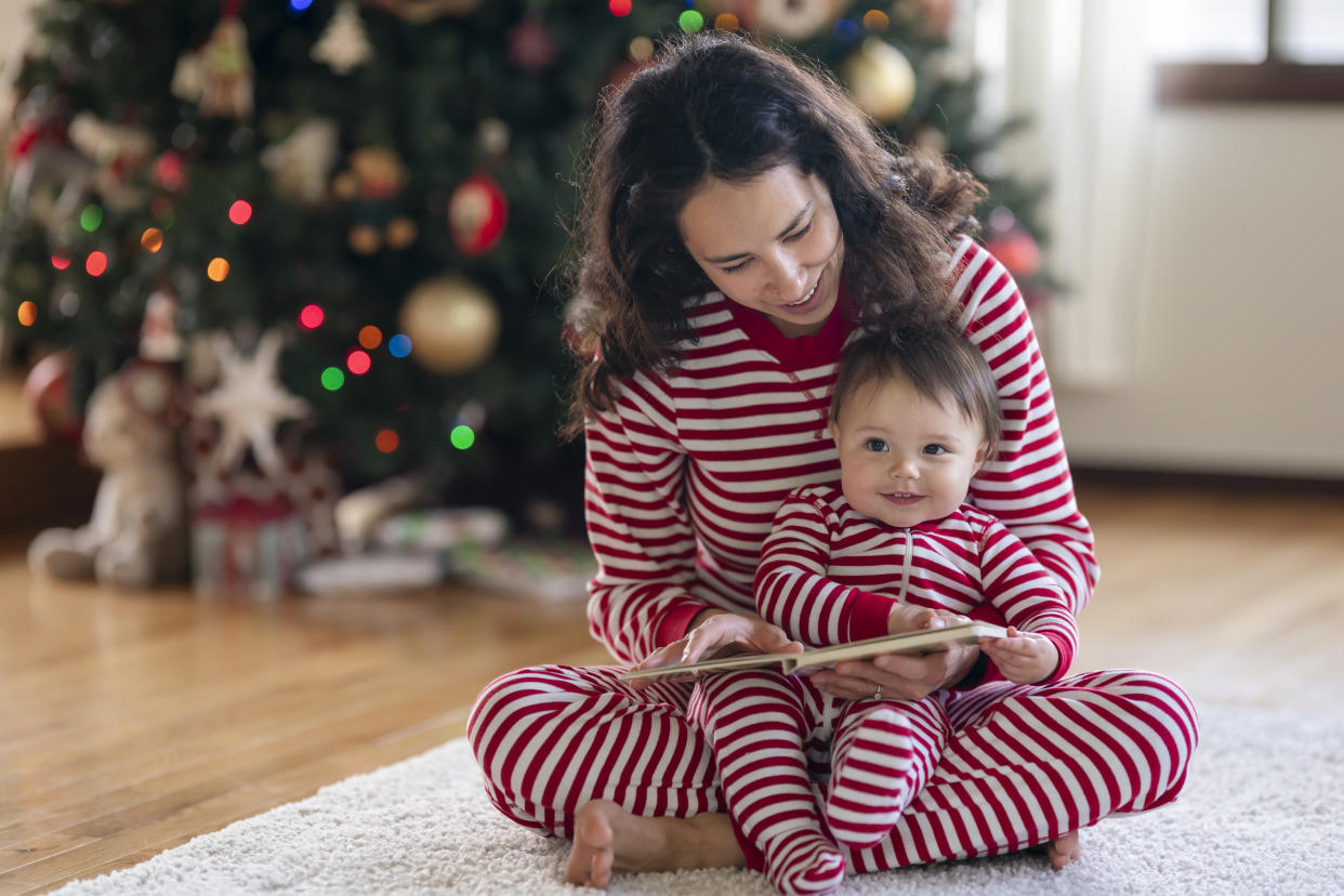 An adorable mixed race baby sits in her mother's lap and smiles as they read a book together. The family is celebrating the baby's first Christmas with a cozy day at home.