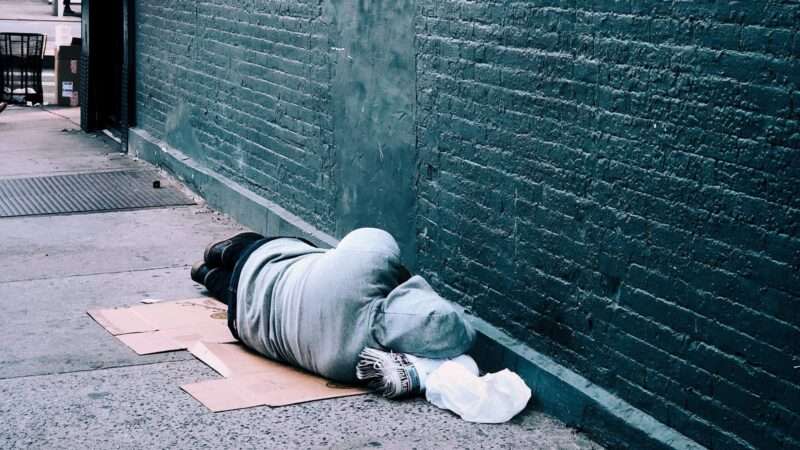 A homeless person sleeps on the street next to a brick wall