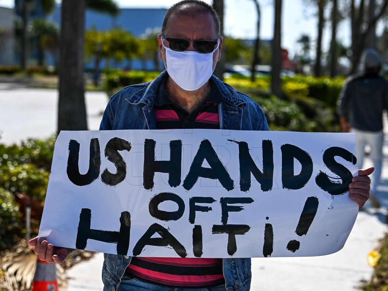 Masked protestor holding up a 'US hands off Haiti!' sign.