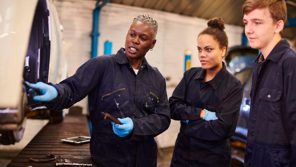 Female mechanic in overalls and blue gloves points something out to a male and a female student also in overalls