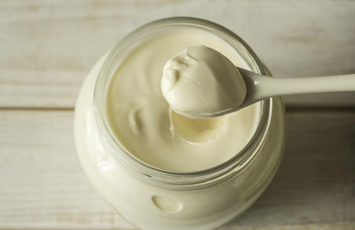 Focus on a spoon with mayonnaise over a full glass jar of mayonnaise on a blurred white wooden table