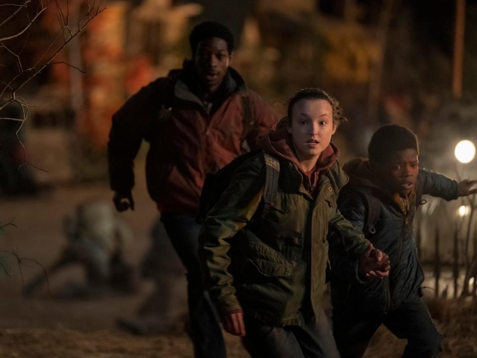 henry, ellie, and sam in episode five of the last of us. they're illuminated by the light of fire, and all three are fearfully running away from something. there are bodies and humans attacking each other in the background