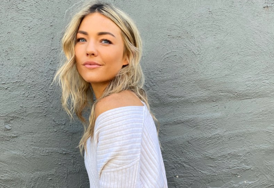 sam frost standing in front of a brick wall wearing a white top