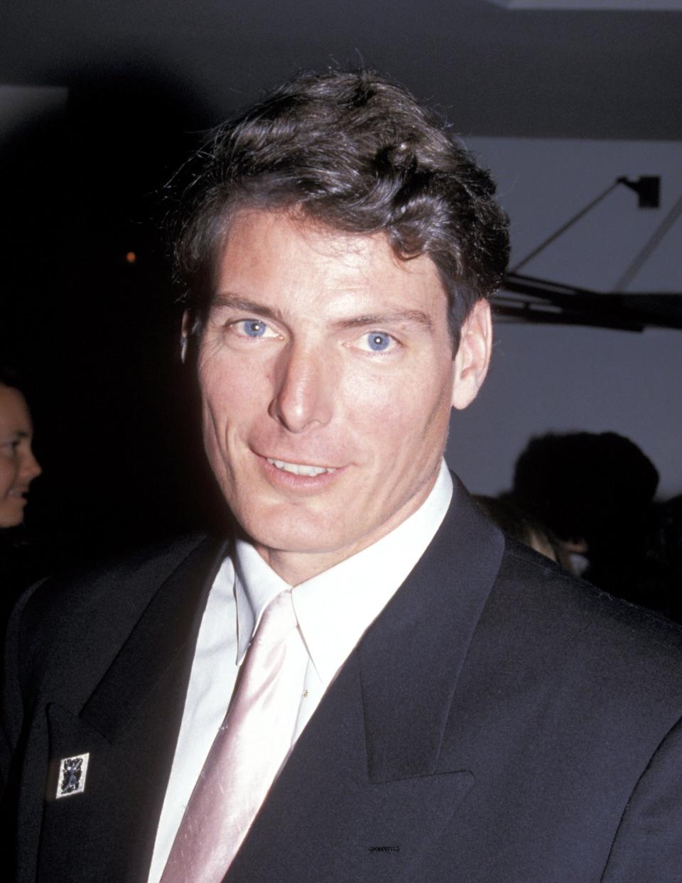 Christopher Reeve, wearing a formal suit with a light-colored tie, smiles for the camera in a public event setting