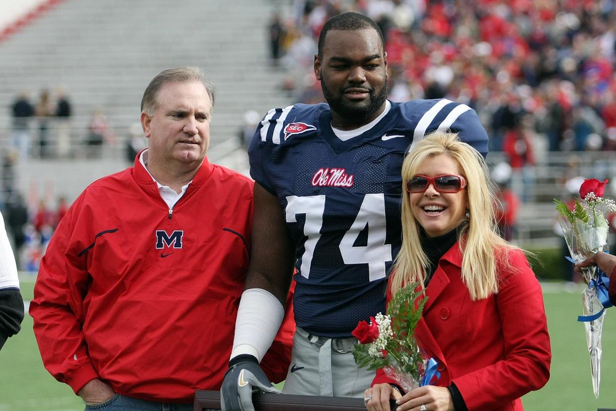 Michael Oher, a Black man who wears a blue Ole Miss Rebels football jersey, stands with his family, a white couple, during a 2008 game.