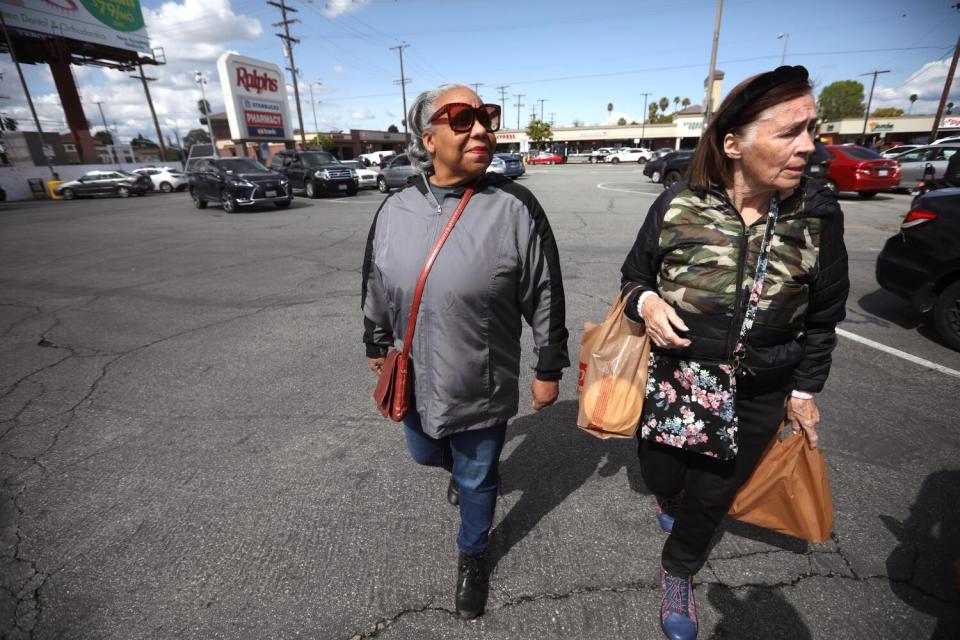 Two older women walk in a parking lot, one carrying grocery bags