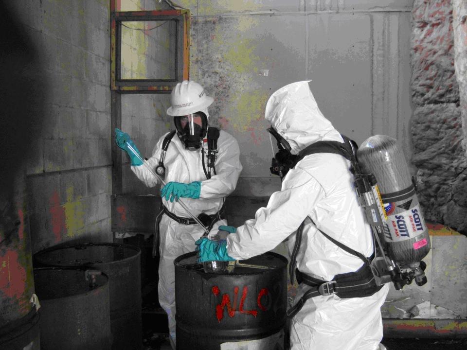 Two men in white protective suits sample a drum of chemicals.