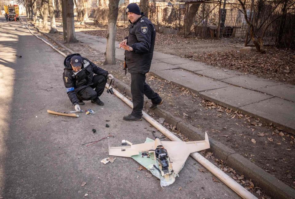 A police officer bends down to look at a part of a broken drone on the ground while one stands and looks at the camera.