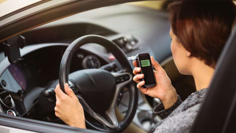 A young woman in the driver's seat of a car looks at a breathalyzer test that reads 0.00