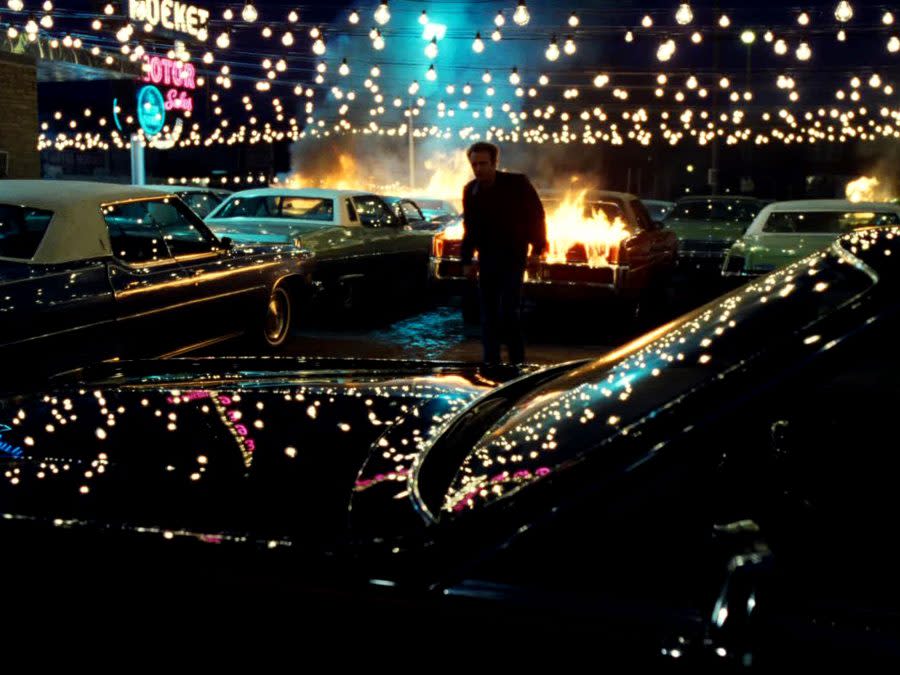 An image from Thief shows James Cann walking away from a fire in a darkly lit car lot