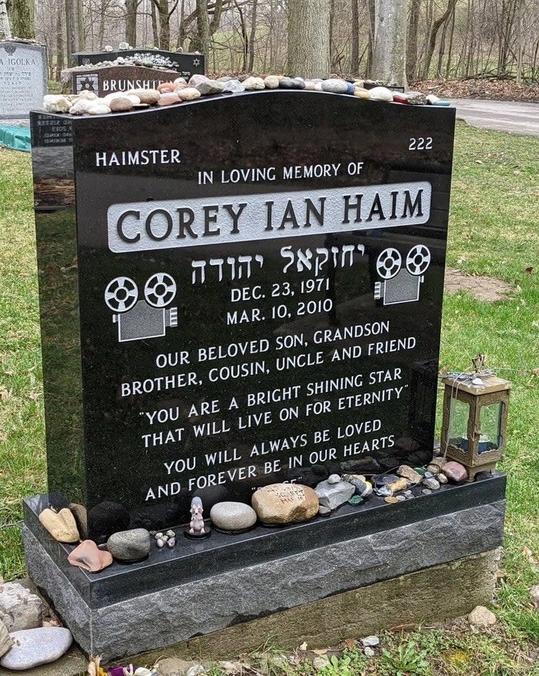 A tombstone with corey's name, film cameras, and the text our beloved son, grandson, brother, cousin, uncle, and friend etched on it