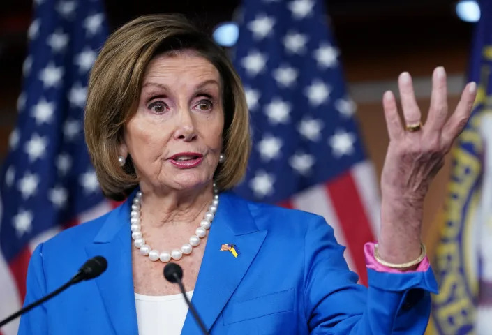 House Speaker Nancy Pelosi, looking indignant and with her hand raised in the air, at the microphone. 