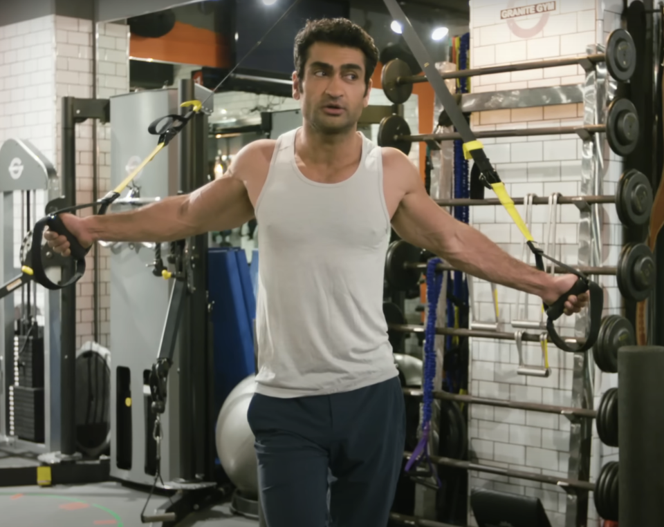 Kumail Nanjiani s'entraîne dans une salle de sport, utilisant des bandes de résistance pour les exercices des bras. Il porte un haut sans manches et un pantalon de sport