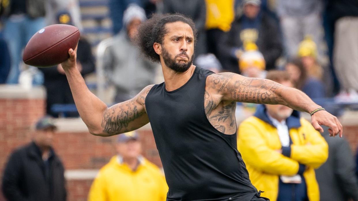 colin kaepernick prepared to throw a football, he wears a black tank top and matching bottoms and has his hair pulled back in a ponytail, spectators are blurry in the background