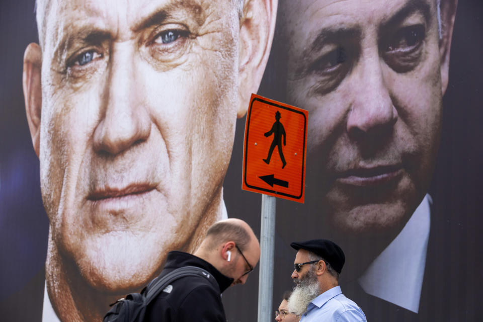 FILE - In this Feb. 23, 2020 file photo, people walk past an election campaign billboard for the Blue and White party, the opposition party led by Benny Gantz, left, in Ramat Gan, Israel. Prime Minister Benjamin Netanyahu of the Likud party is pictured at right. Netanyahu and Gantz announced Monday, April 20, 2020, that they have forged a deal to form an “emergency” government. The deal between the Likud Party and the Blue and White ends months of political paralysis and averts what would have been a fourth consecutive election in just over a year. Terms of the agreement weren’t immediately announced. (AP Photo/Oded Balilty, File)