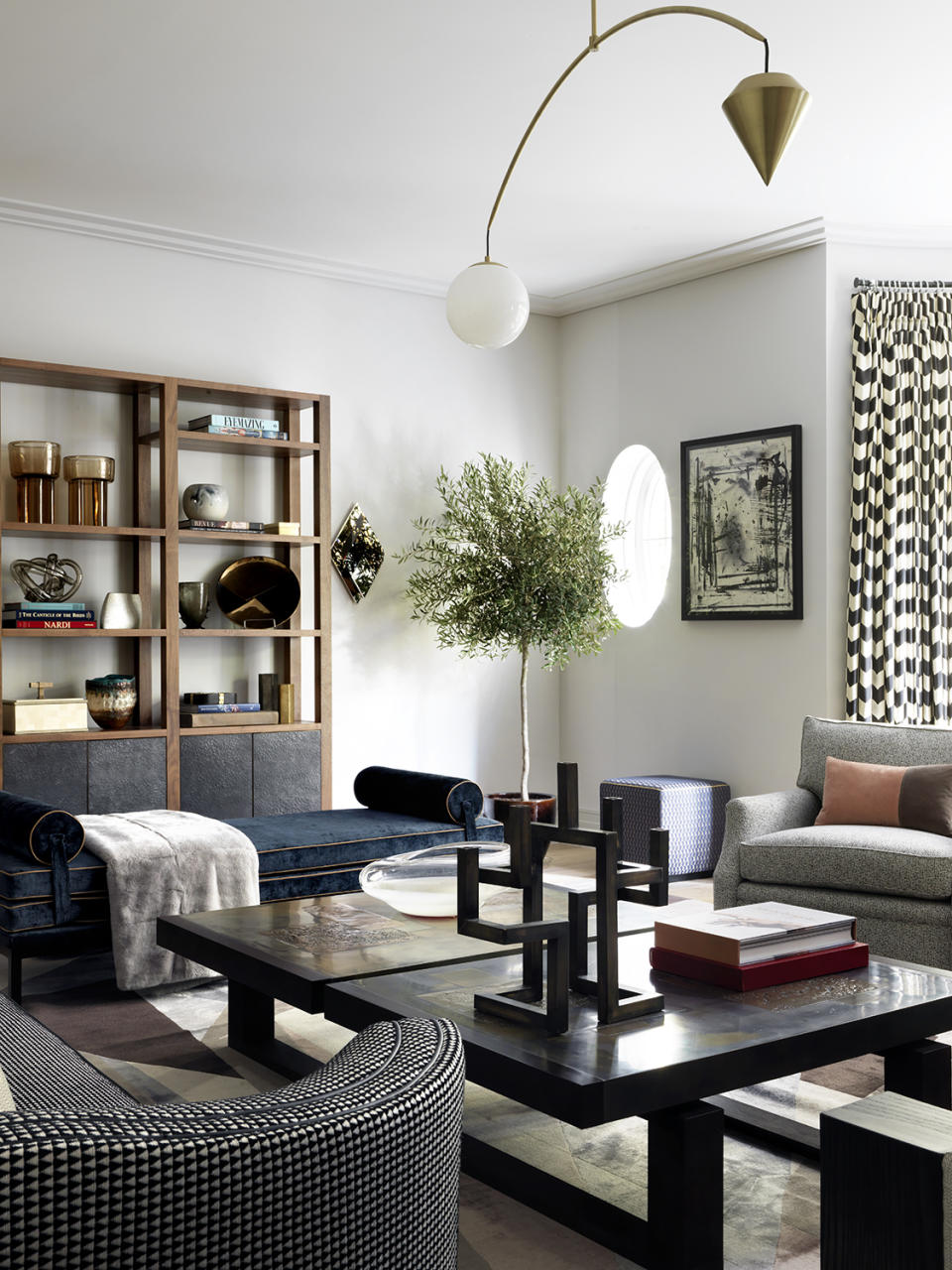 Modern living room with striking geometric ceiling light, black coffee table, two armchairs and blue sofa, houseplant, wooden shelving unit, round window, artwork on wall.