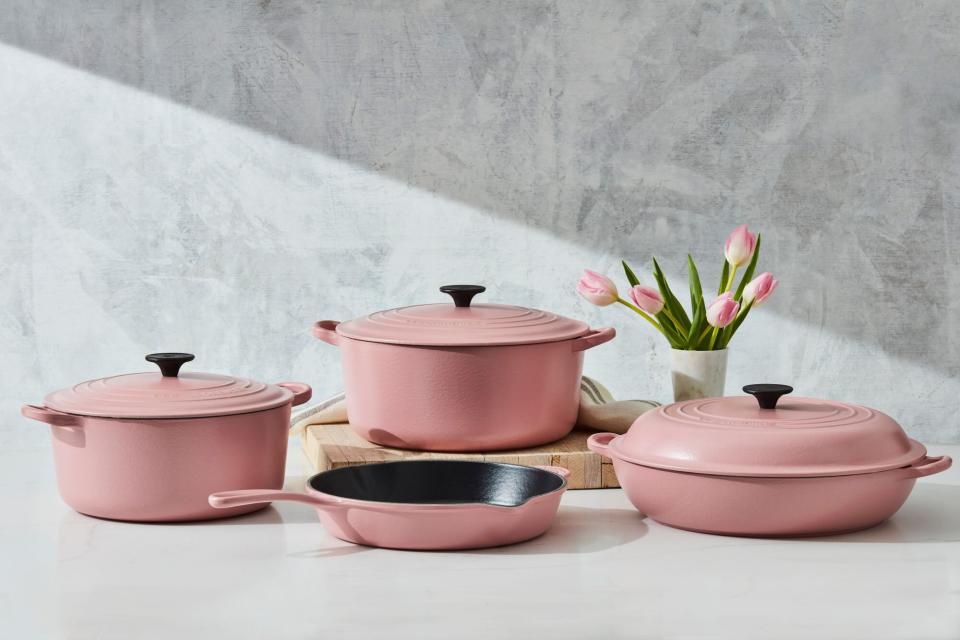 two baby pink pots with lids, one braiser with a lid and a deep skillet pictures on a white countertop with a white butcher block and a vase of pink tulips