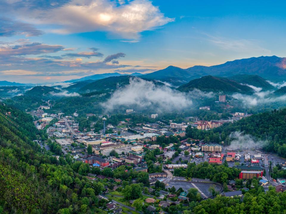 A view of the town of Gatlinburg, Tennessee surrounded by mountains, trees, and with floating clouds.