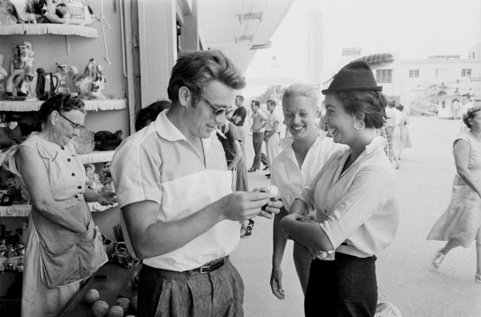 James Dean wearing sunglasses, interacts with Ursula Andress and Gwen Verdon outside a shop. Dean and Verdon are smiling while Andress looks towards Dean