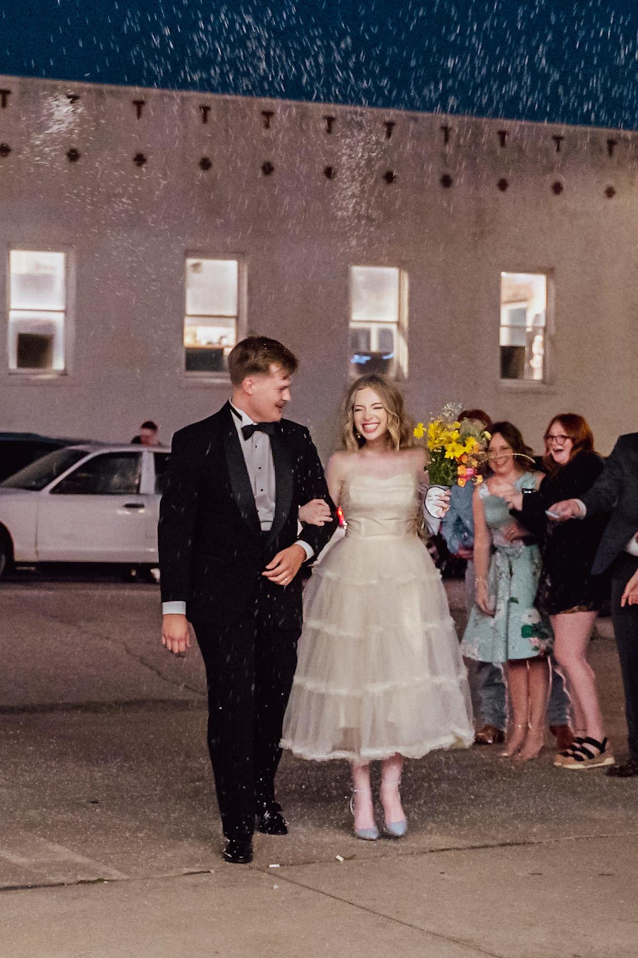 A bride and groom grin and walk out of their wedding reception together.