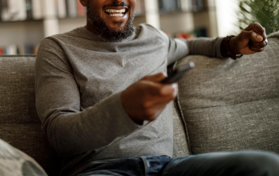 A man with a beard and a big smile sits on a sofa, holding a remote control in his right hand, watching TV