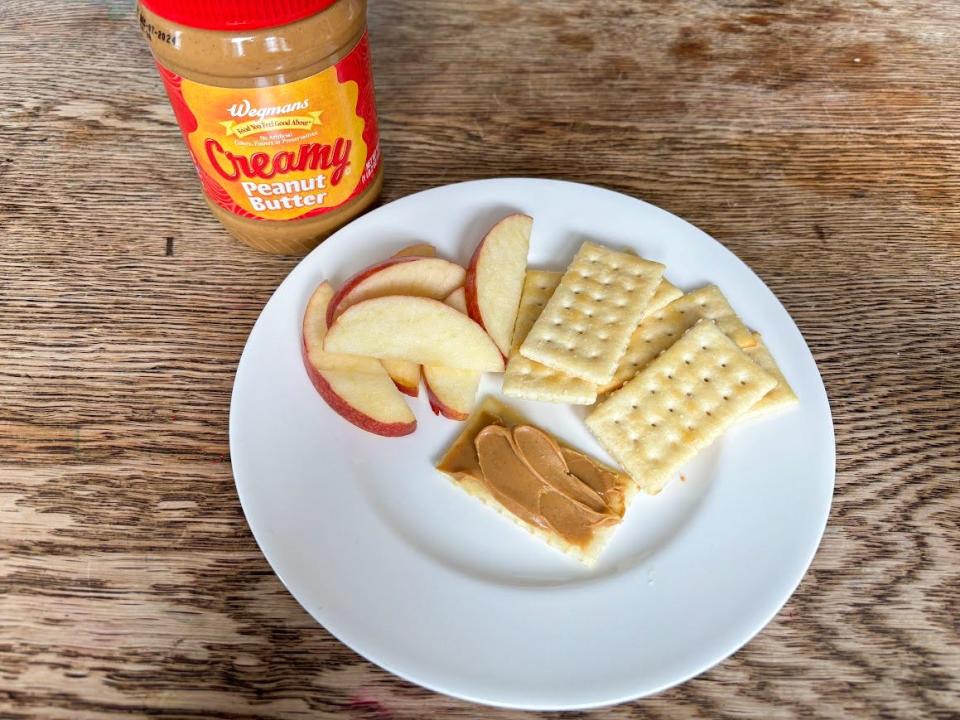 A jar of peanut butter with an orange and red label sits next to a plate of apple slices and crackers. Thick-looking peanut butter is spread on one cracker