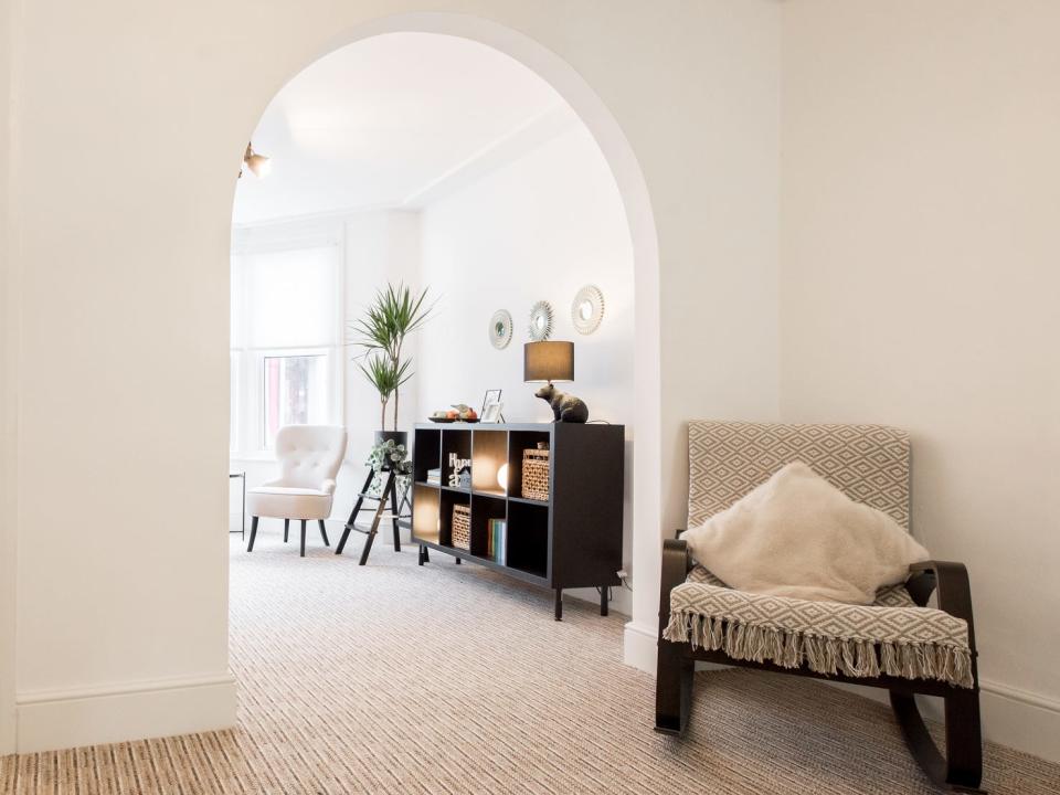 Home with a white walls, a rocking chair with a beige and white blanket, and an archway leading to living room with entertainment center, a plant, and a white chair