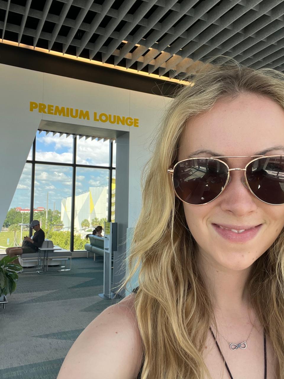 A woman with blonde hair and aviator sunglasses poses in front of a sign that reads premium lounge.