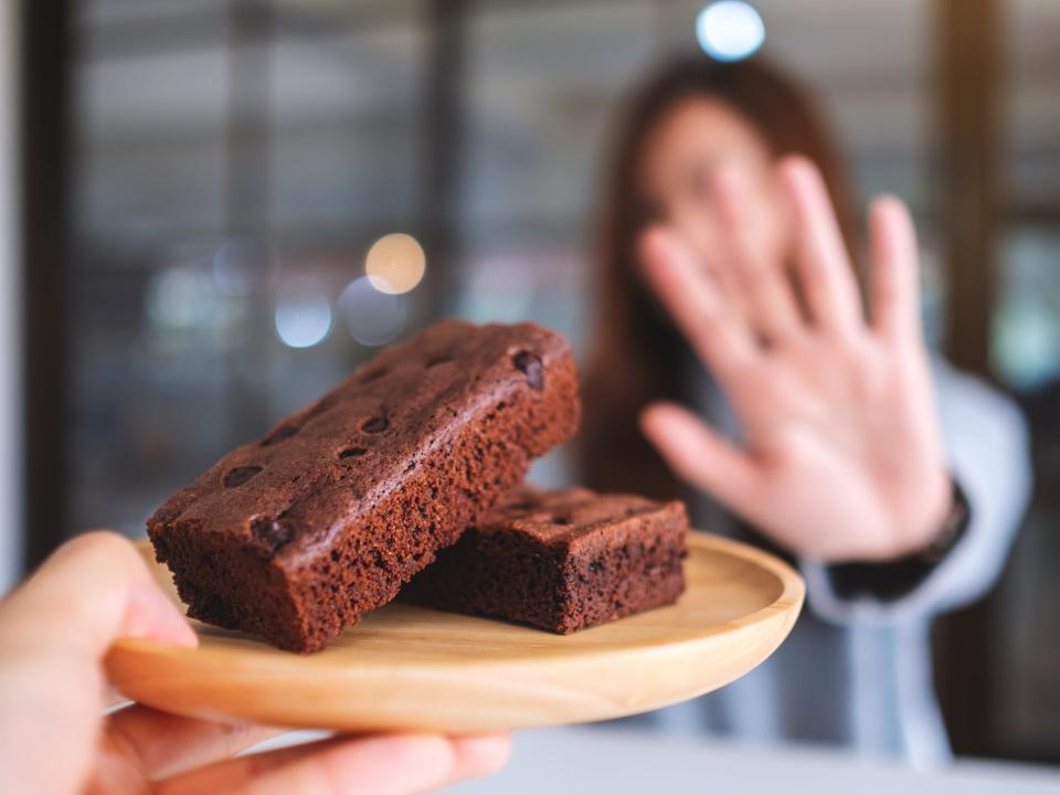 a woman pictured out of focus in the background, putting her hand out in a 'stop' signal to refuse a plate of chocolate brownies, saying no to dessert