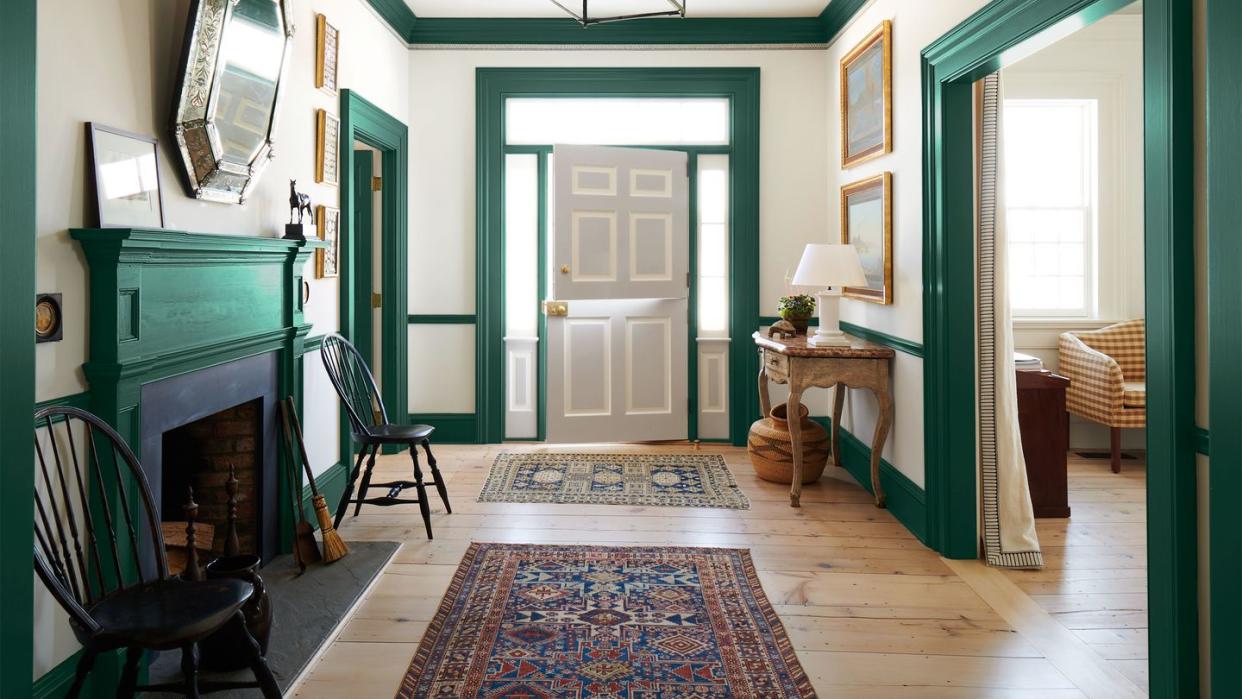 an entry hall has off white walls with rich green moldings and woodwork, a dutch door painted light gray, a fireplace, two black wooden chair, two caucasian rugs, a marble topped half moon console, and a gallery lantern