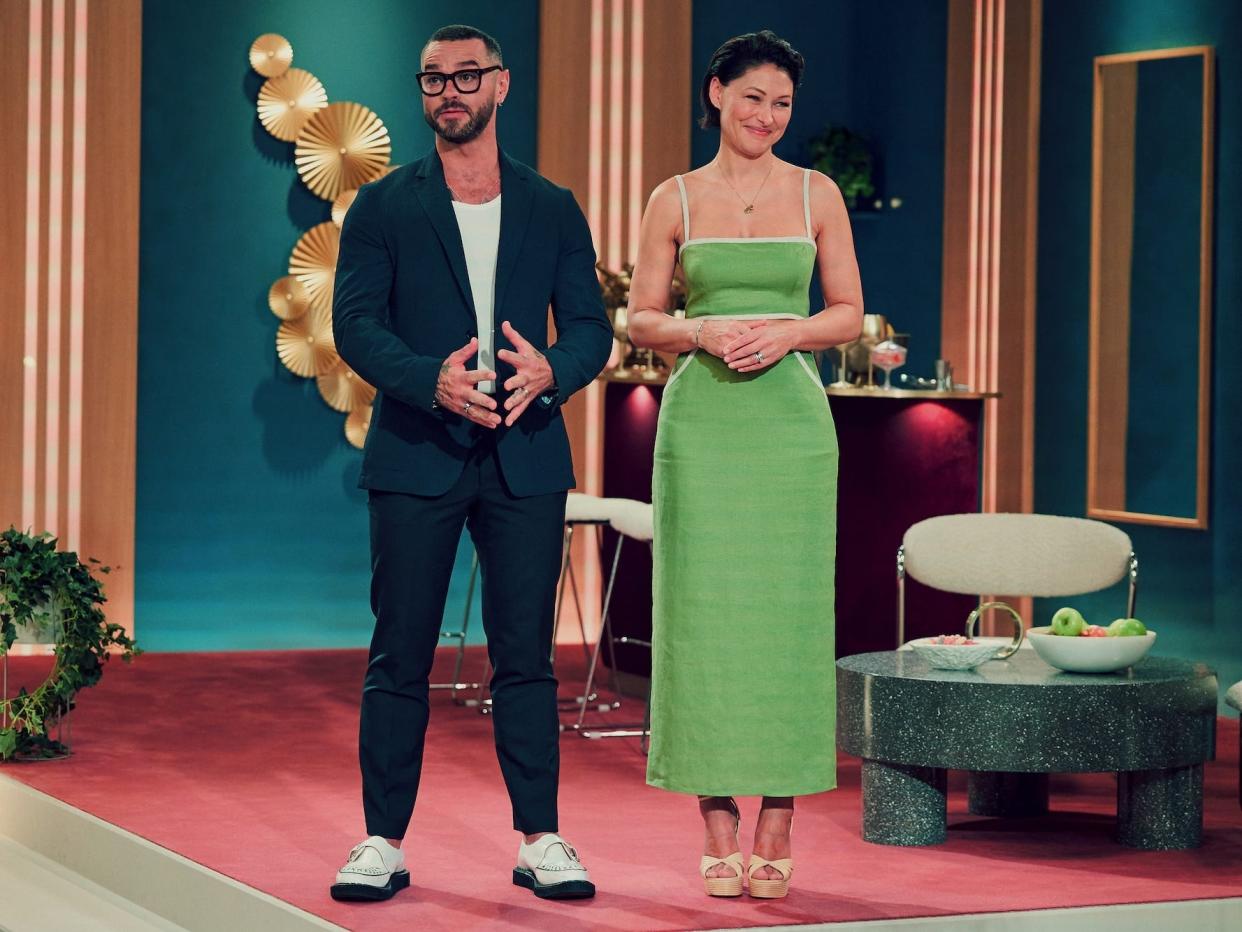 A still from "Love Is Blind: UK" featuring Matt Willis with a blue suit standing next to Emma Willis in a green dress in a lavish room.