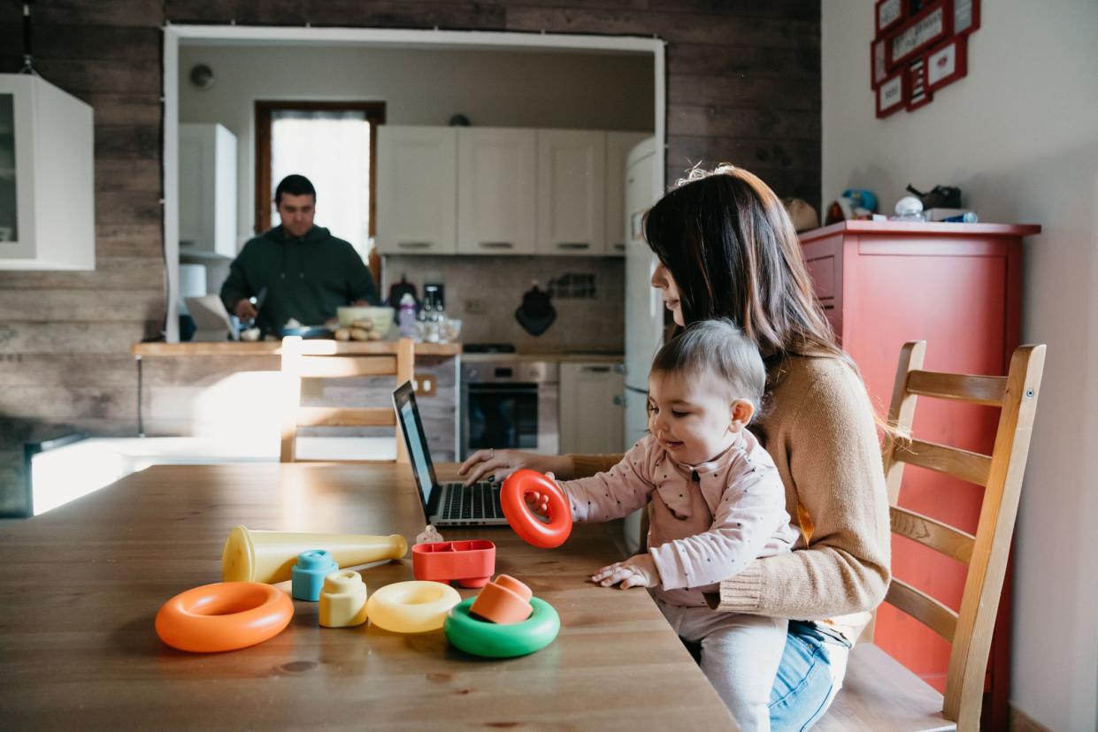 A little girl using a laptop with her mom while the dad is cooking in the kitchen. They are sitting at the table, the mother is holding her daughter. Family locked down during Coronavirus Covid-19 quarantine.