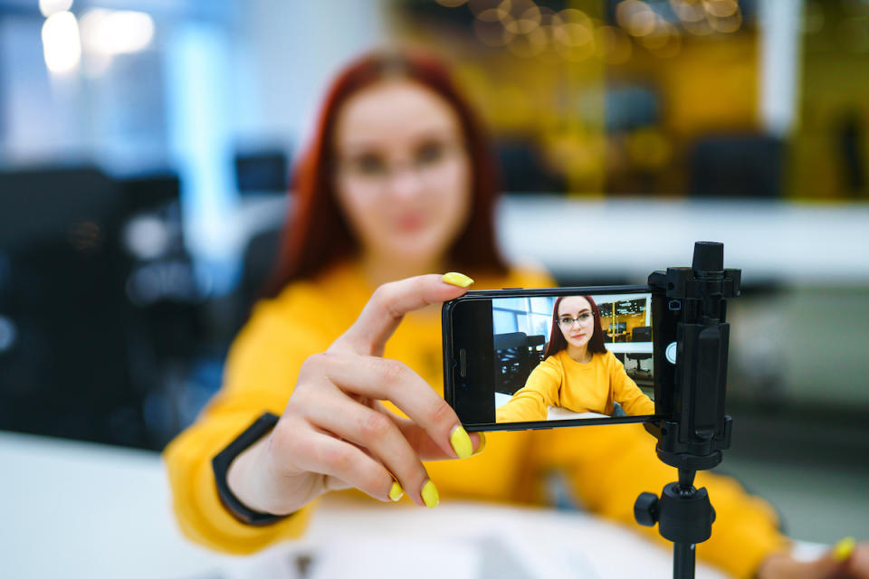 Hand woman holding camera for recording video while sitting at office. Teenager student in a yellow sweater with laptop having fun vlogging live feeds on social media. Technology and videoblog concept