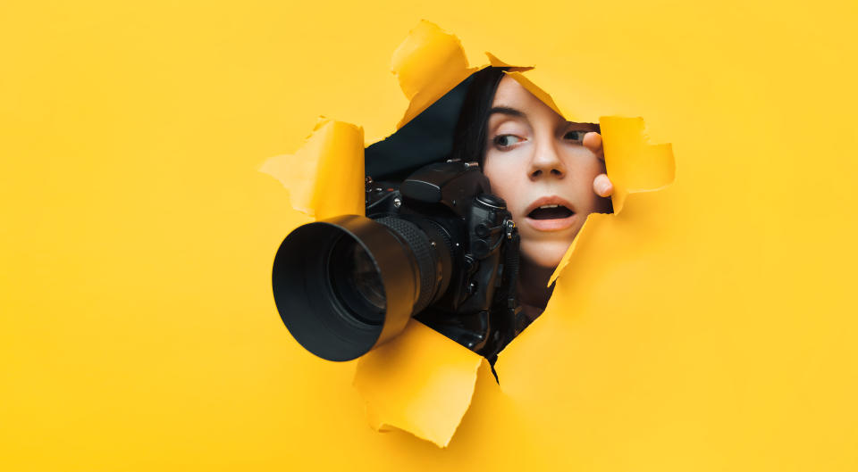 A young paparazzi girl with a reflex camera looks out from cover and looks at what is happening with her mouth open. Yellow paper, torn hole. Tabloid press. In search of the plot for photo stocks.