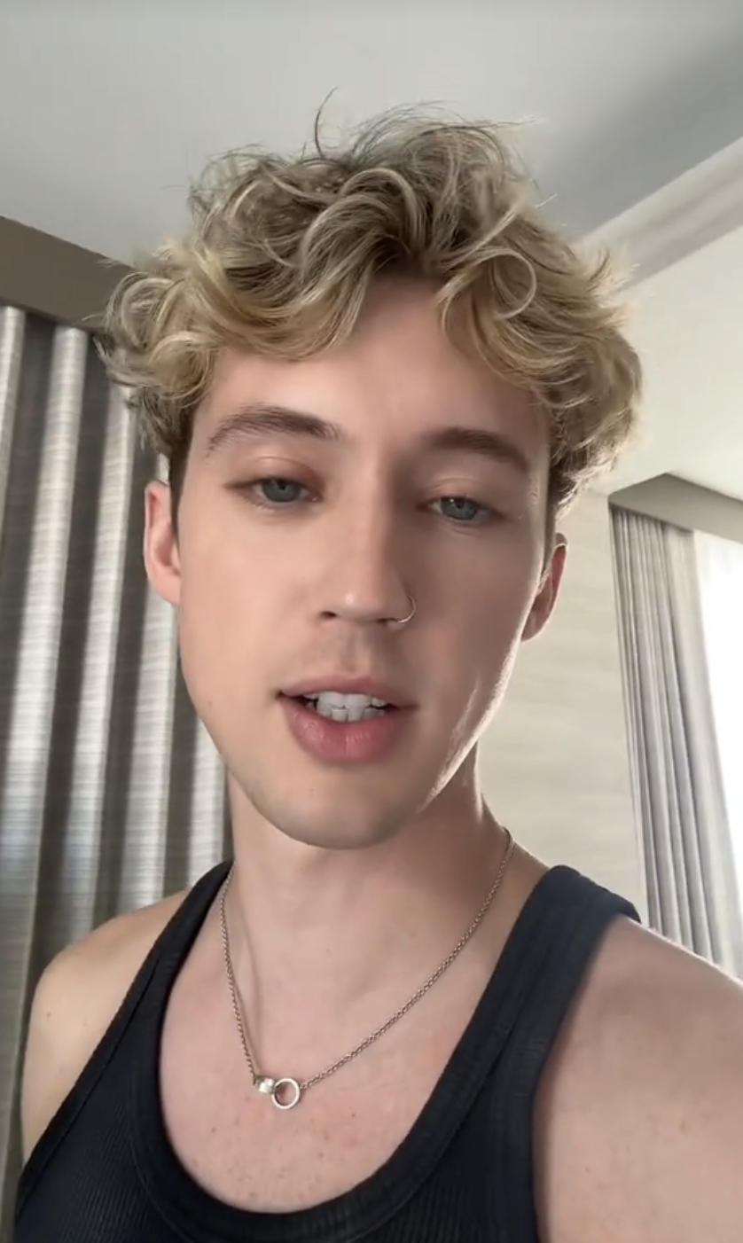 Troye Sivan wearing a tank top and necklace, standing indoors with light streaming through curtains in the background