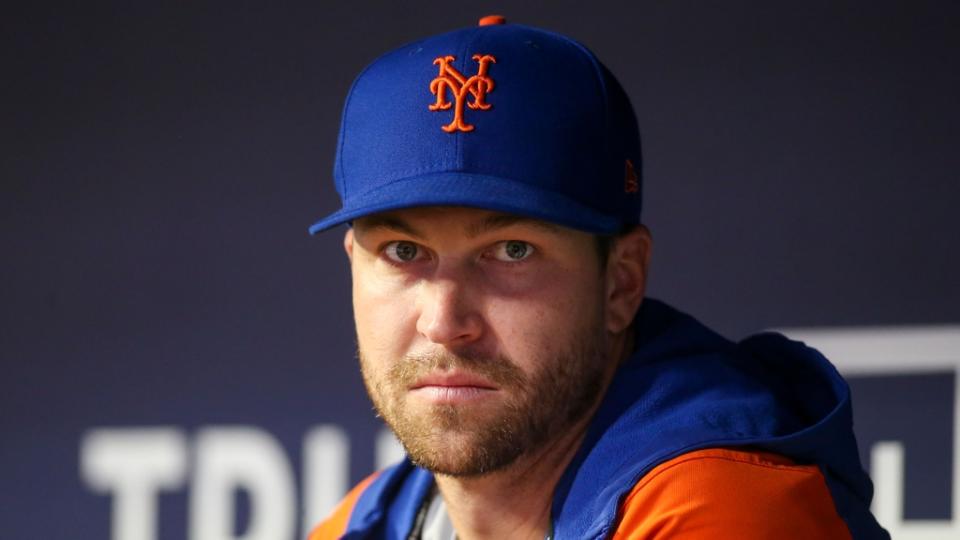 Jacob deGrom looking into camera, sitting in dugout wearing hoodie and hat