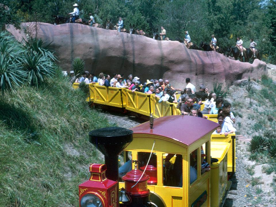 A yellow and red open train style ride with people sitting in it and a ledge overhead with peopling riding donkeys.