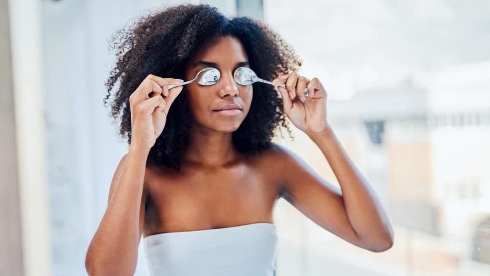 A woman in a white top holding two cold spoons over her swollen eyelids