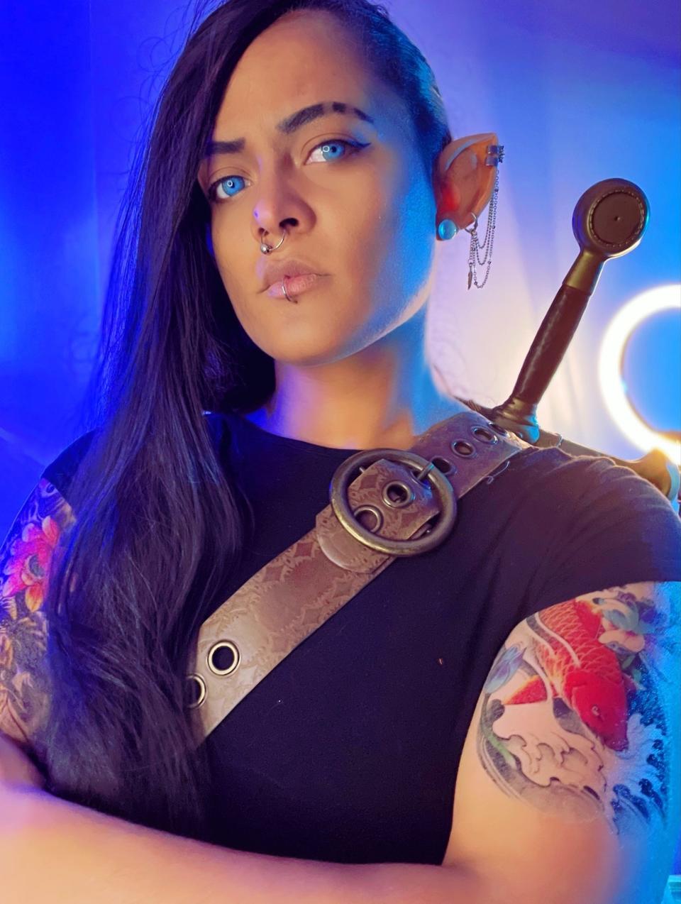 A person with long, black hair and blue eyes poses with a sword.