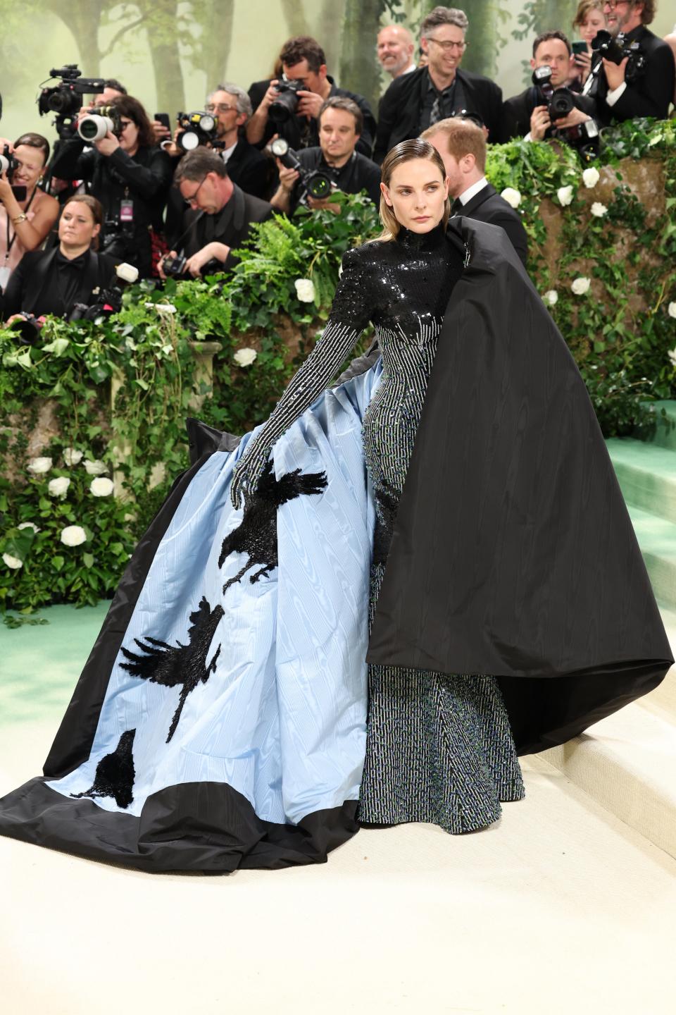Rebecca Ferguson in a gown with a blue skirt featuring bird designs and a black cape, posing at an event with photographers in the background