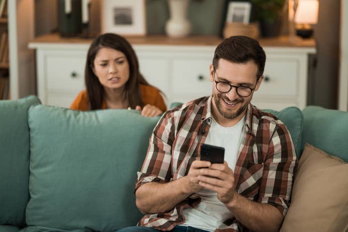 Man wearing glasses and a plaid shirt smiles while using his phone on a couch. A woman behind him looks upset and leans over the couch, peering at the phone