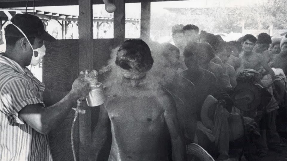 This photo taken in Hidalgo, Texas, in 1956 shows a masked worker spraying <em>braceros</em> with DDT while others wait in line. - USCIS History Office and Library/Department of Homeland Security