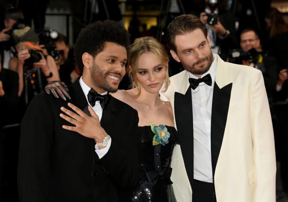 the weeknd, wearing a black tuxedo, lily rose depp, wearing a green dress, and sam levinson, wearing a white tuxedo, posing for a photo together as photographers take pictures behind them