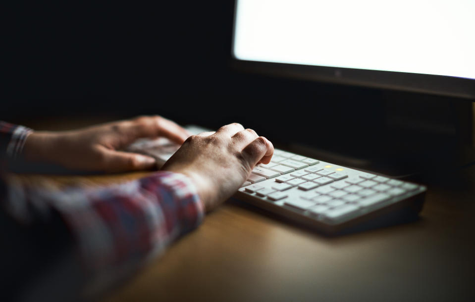 Hands type on a computer keyboard in front of the PC monitor whose light is providing the only illumination.