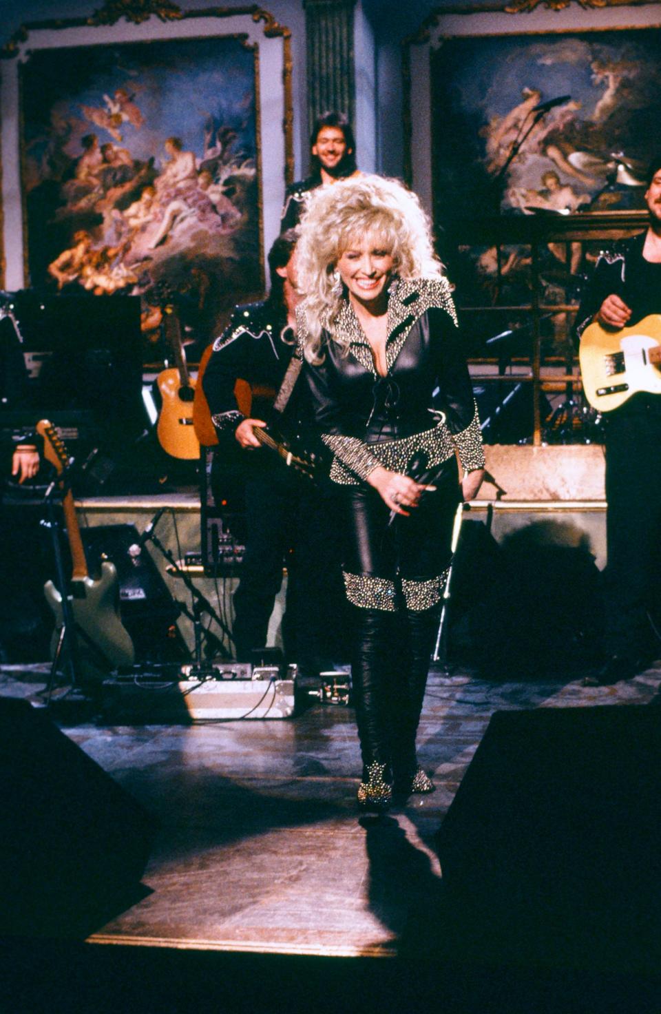 Dolly Parton singing in a fitted black leather jumpsuit with gold studded accents on the collar, shoulders, sleeve cuffs, belt, and knees.