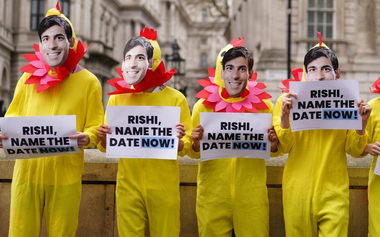 A row of seven people in cheap yellow chicken suits carrying placards asking Sunak to name a date, and wearing Sunak masks.