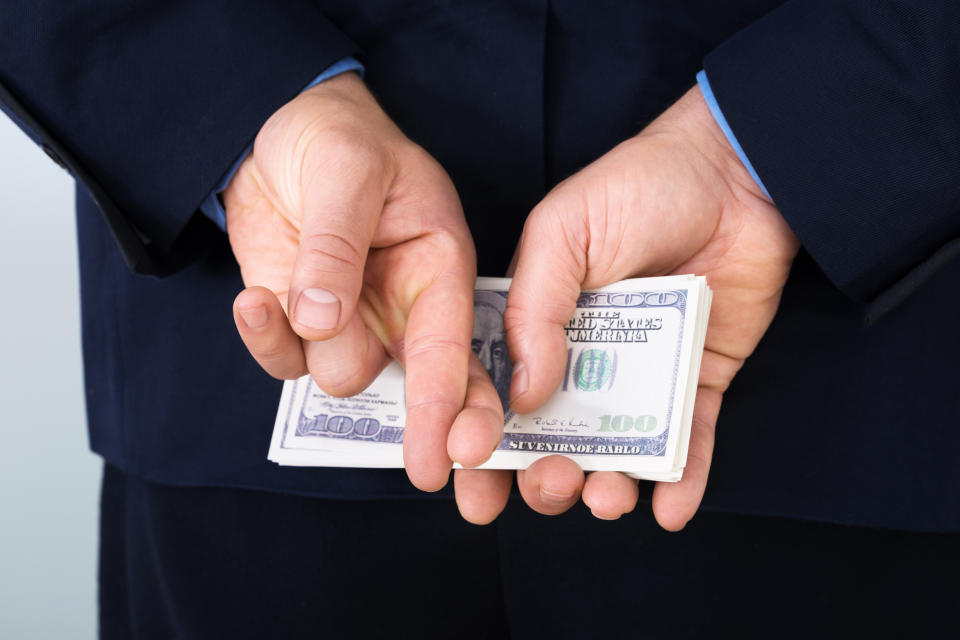 A businessman holding a stack of hundred-dollar bills behind his back, while also crossing his fingers.