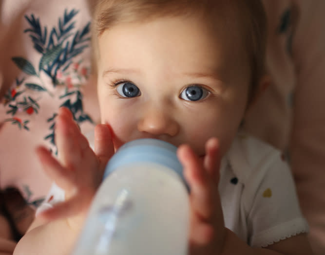 A baby with blue eyes holds a baby bottle with both hands and looks at the camera. There's a patterned fabric in the background